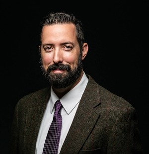 professor wearing a black suit and white button up shirt with a black tie in front of a black backdrop